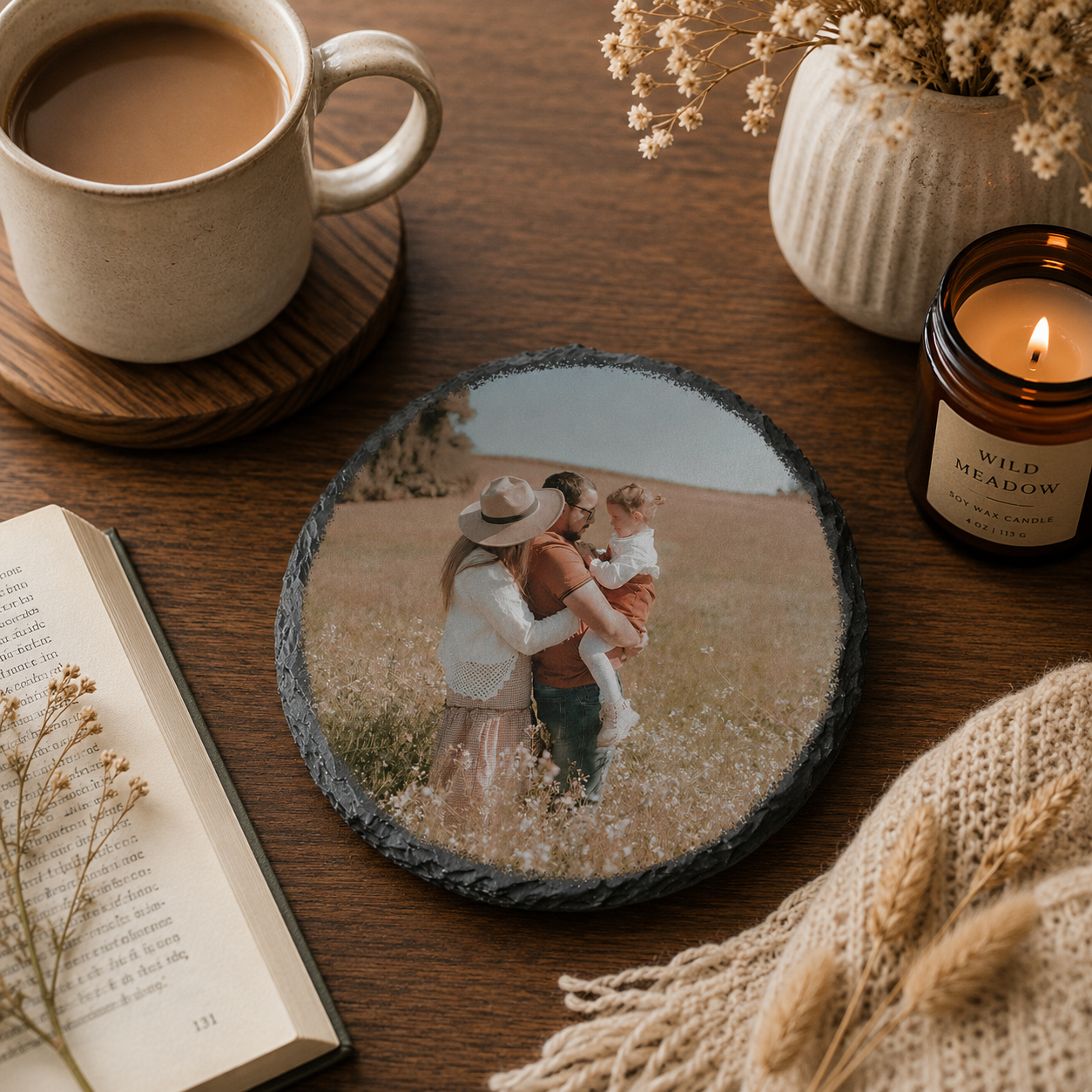 Round slate coaster with a family photo of a couple and child on a wooden table with a cup of coffee, book, and candle.