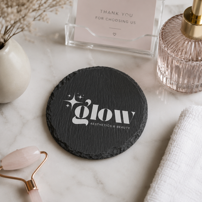 A round black slate coaster with a personalised custom logo in white, styled on a white marble surface with spa props including a rose quartz roller, a folded white towel, a glass bottle, and a card holder.