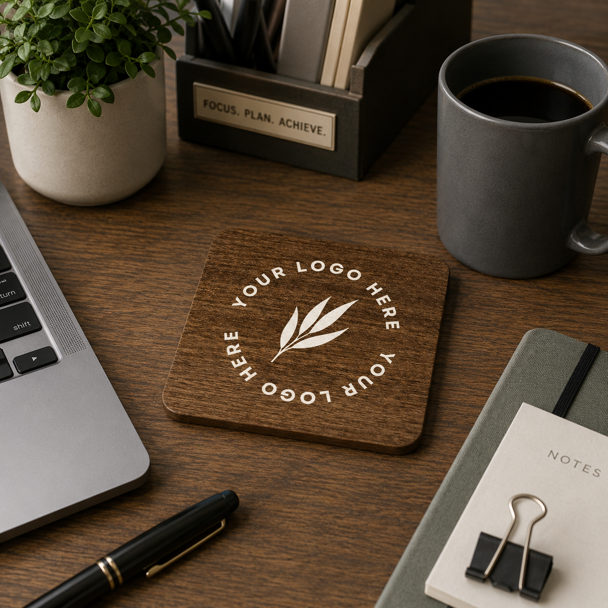 A square wooden coaster with a custom logo design featuring a leaf motif, displayed on a wooden desk surrounded by a laptop, coffee mug, notebook, and pen.