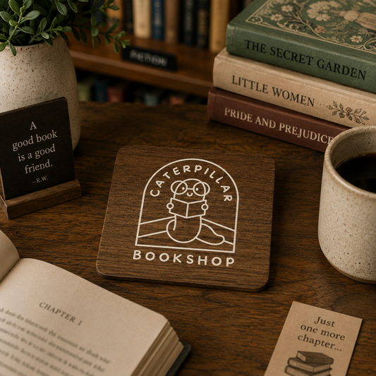 A square wooden coaster with a white bookshop logo sits on a wooden table surrounded by books, a mug of coffee, and a small sign.