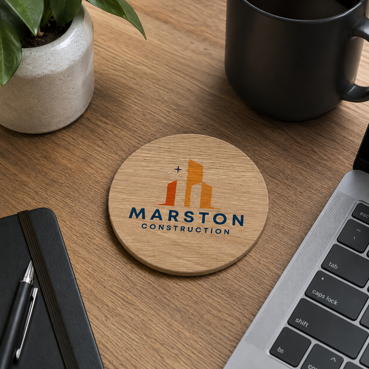 A round wooden coaster with a custom business logo in navy blue and orange, displayed on a wooden desk surrounded by a laptop, notebook, plant, and a black mug.